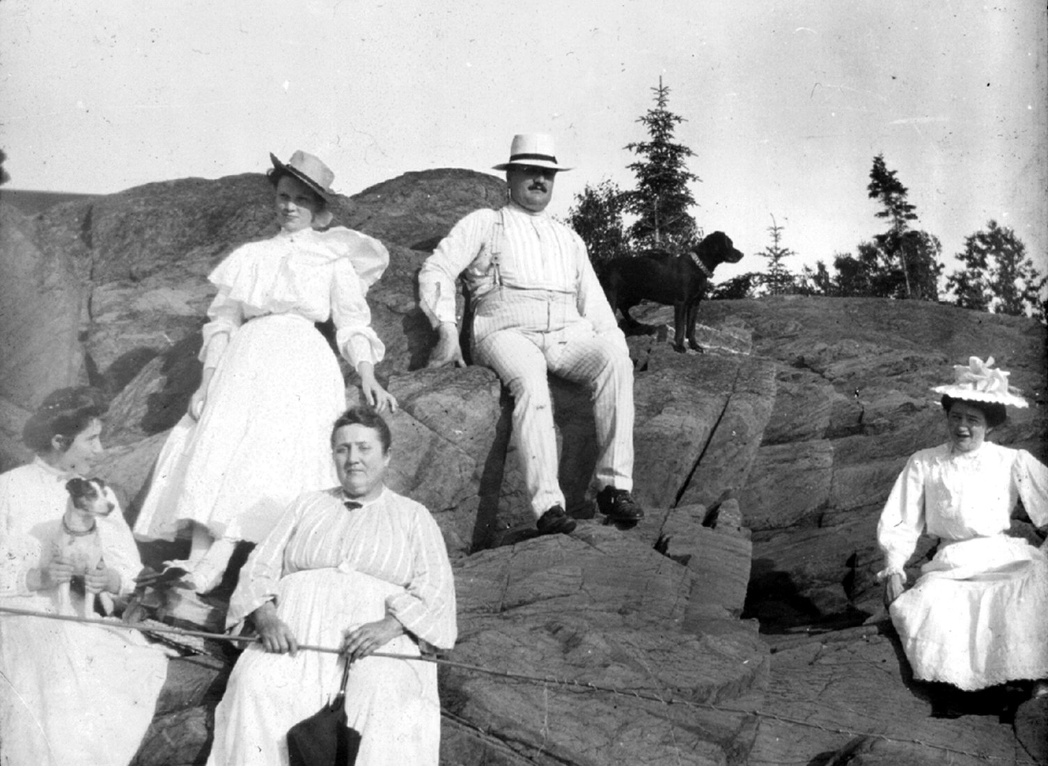 Five adults wearing white and sitting on a rock, with two dogs. One of the women is holding a fishing rod.