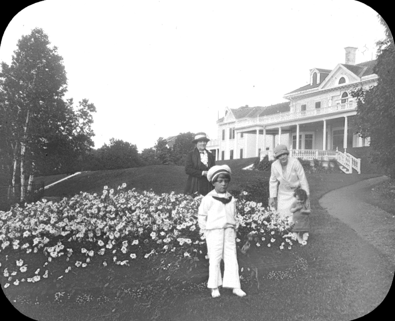 Two women and two children near flowerbeds in front of a very posh residence.