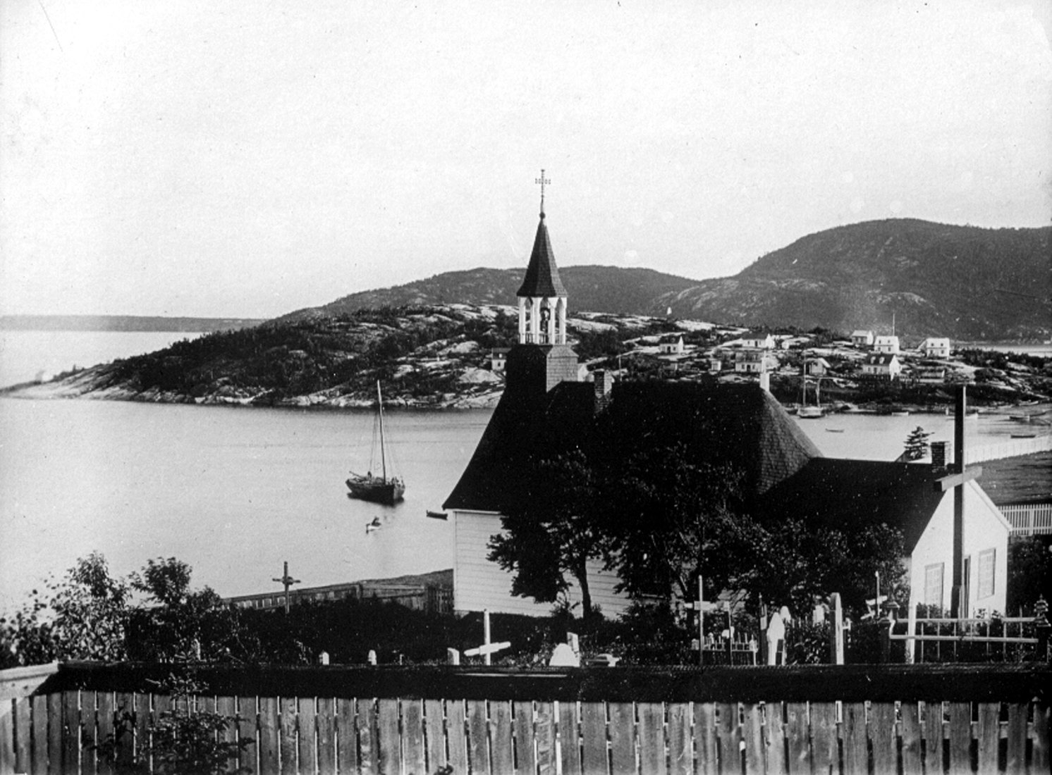 Une chapelle et son cimetière surplombent une baie, où sont amarrées des embarcations, dont deux bateaux à voile.