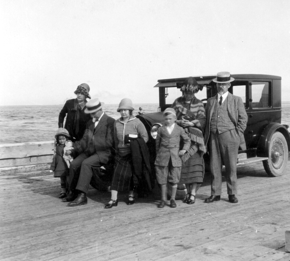 Five adults and two children on vacation, posing in front of an old car parked on a wharf.