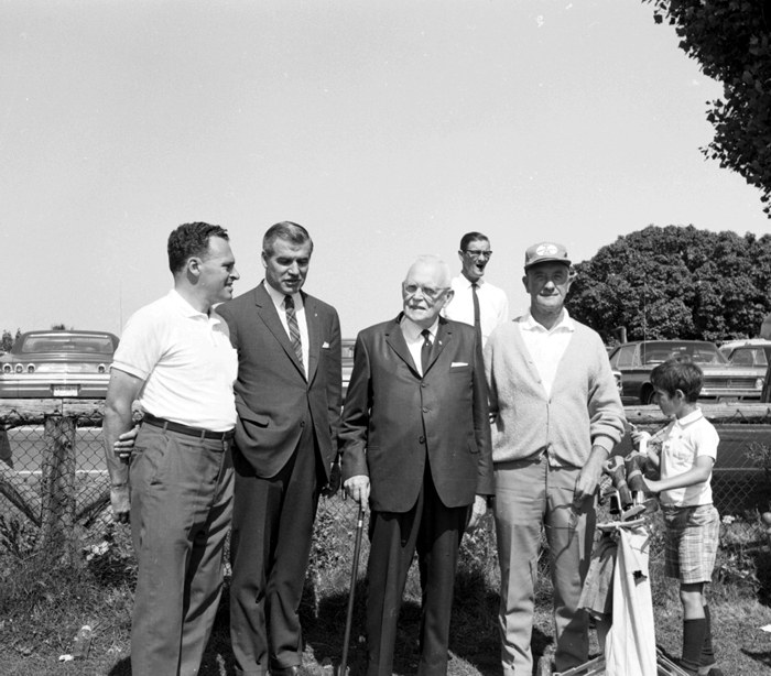 Portrait of a group of four men, followed by a caddie carrying a golf bag, with a parking lot in the background.