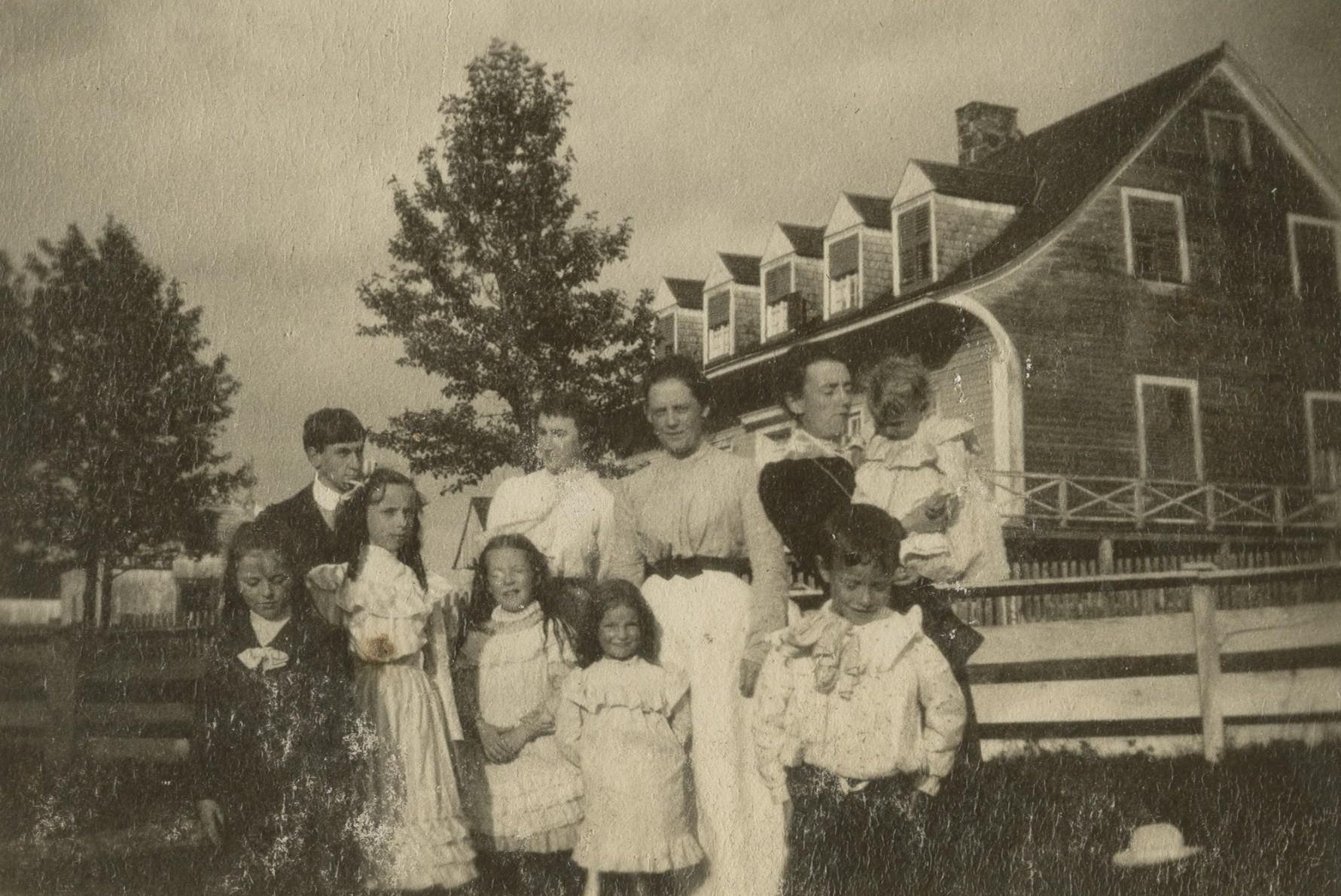 Three women and several children posing in front of a large old house with cedar shingling.