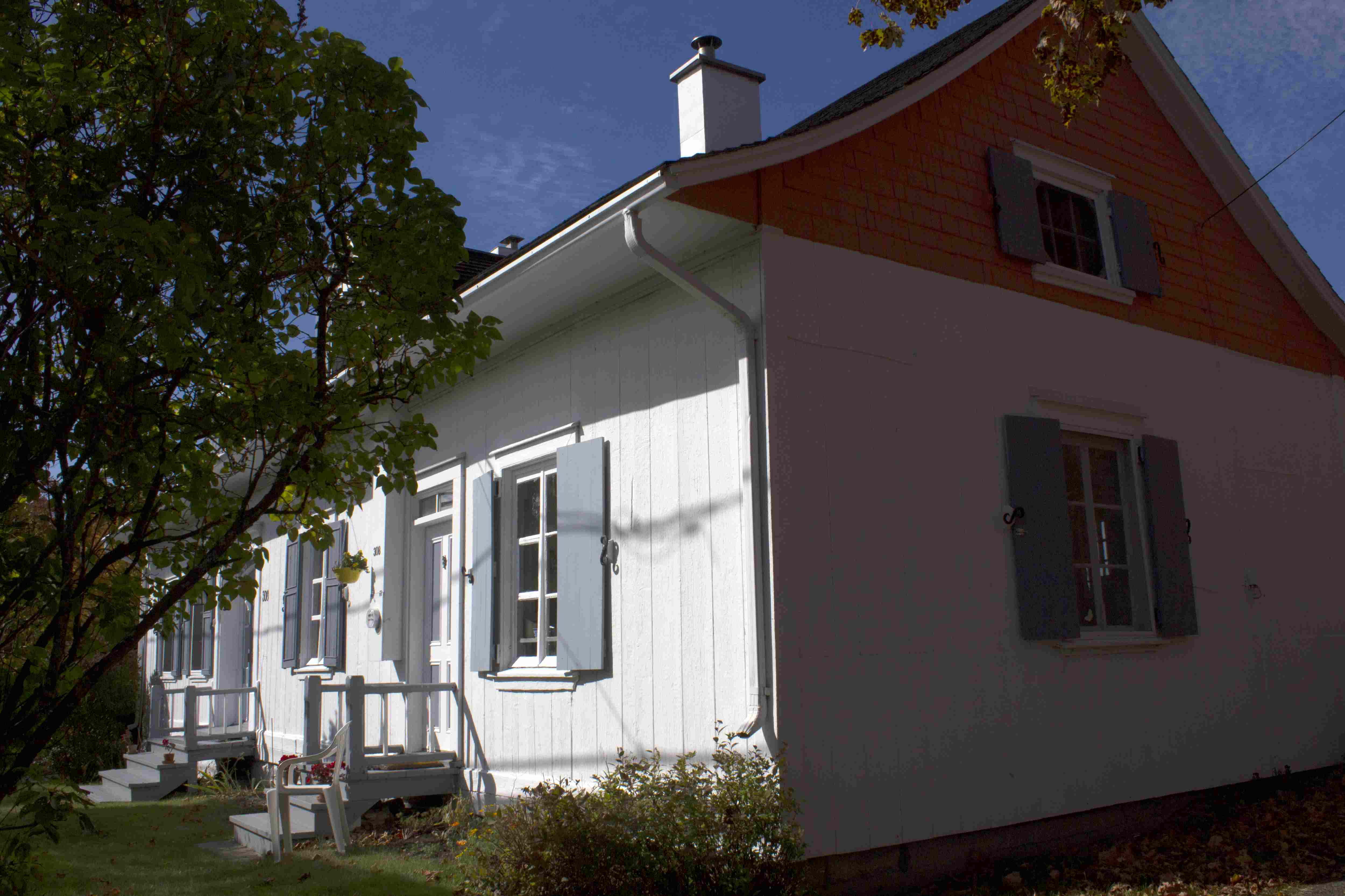 Photographie en couleurs d’une maison ancienne blanche aux volets bleu pâle, entourée de verdure.