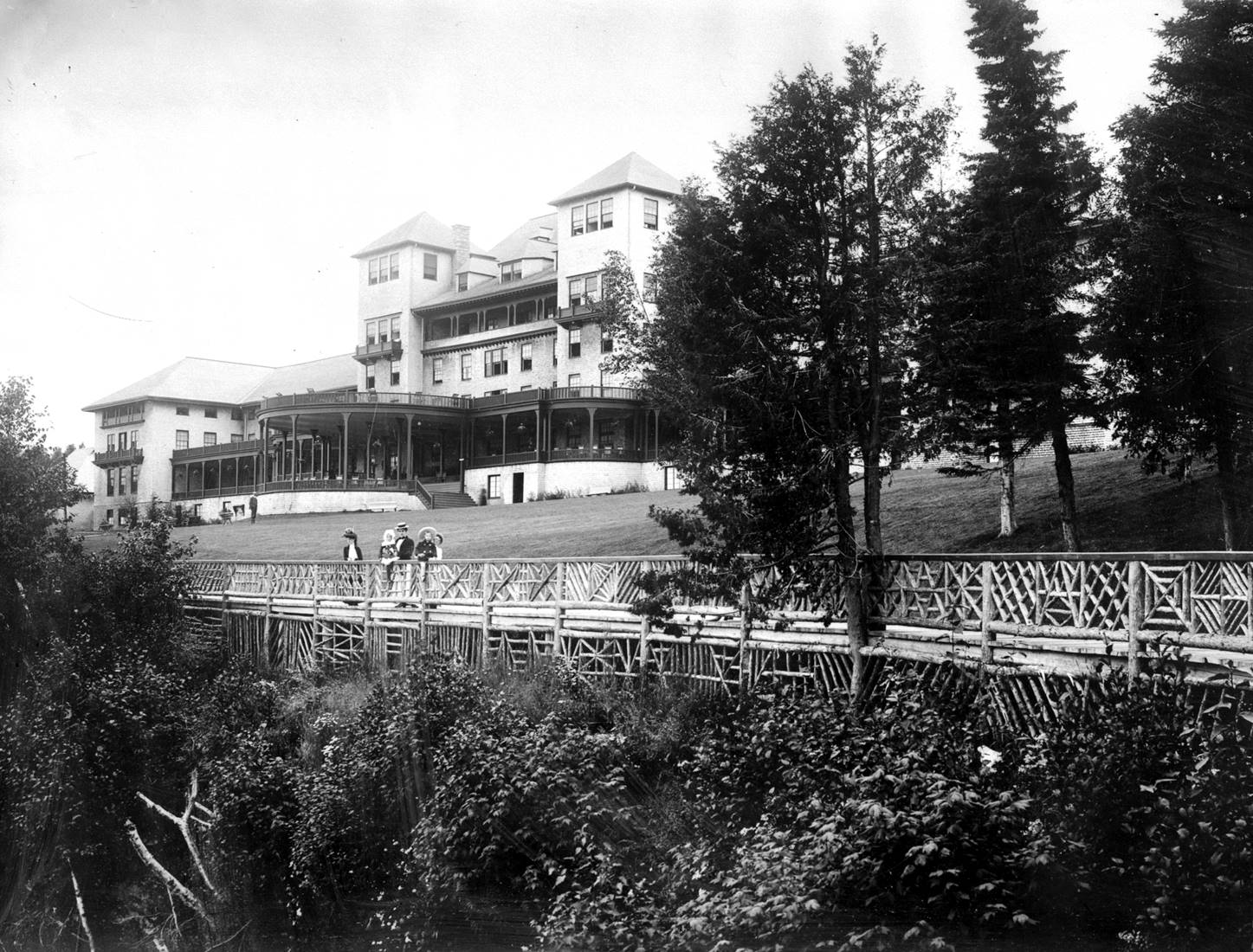 A large hotel with big verandas, ladies and young childrentaking in the scenery near a fence.