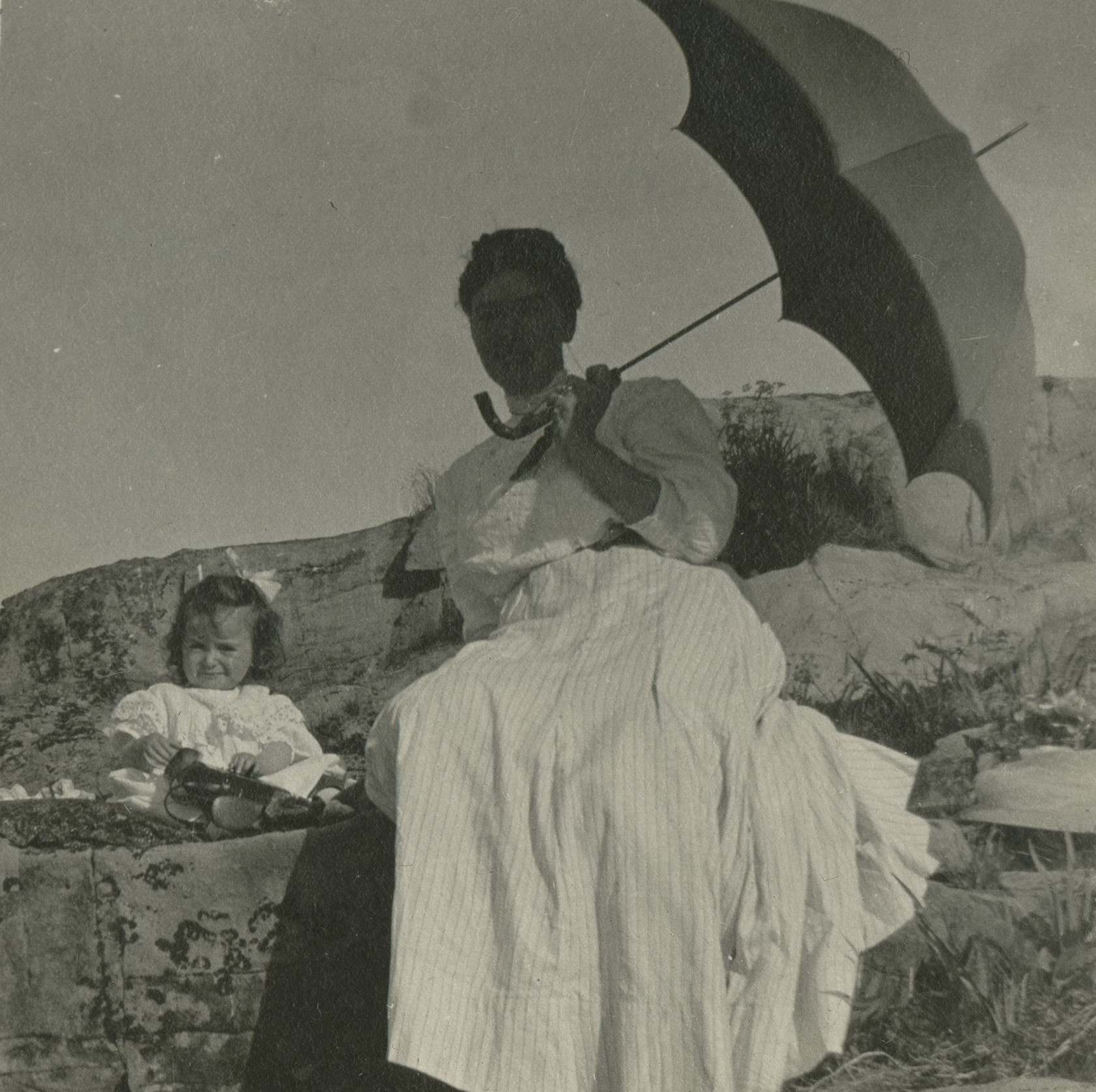 A women in a long dress and a young child sitting on a rock, with a parasol protecting them from the sun.