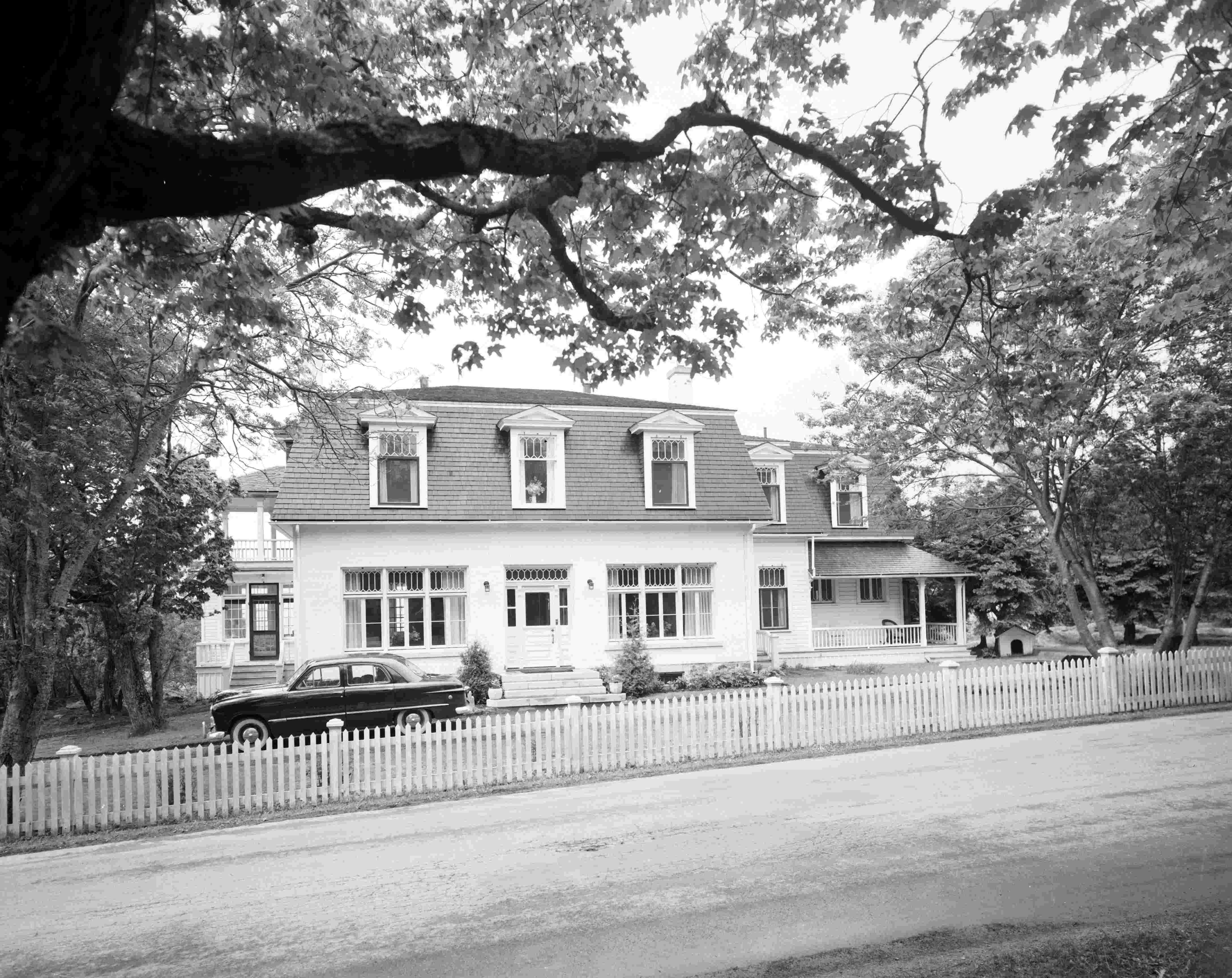 A white house with a mansard roof near a dirt road, with an automobile parked out front.