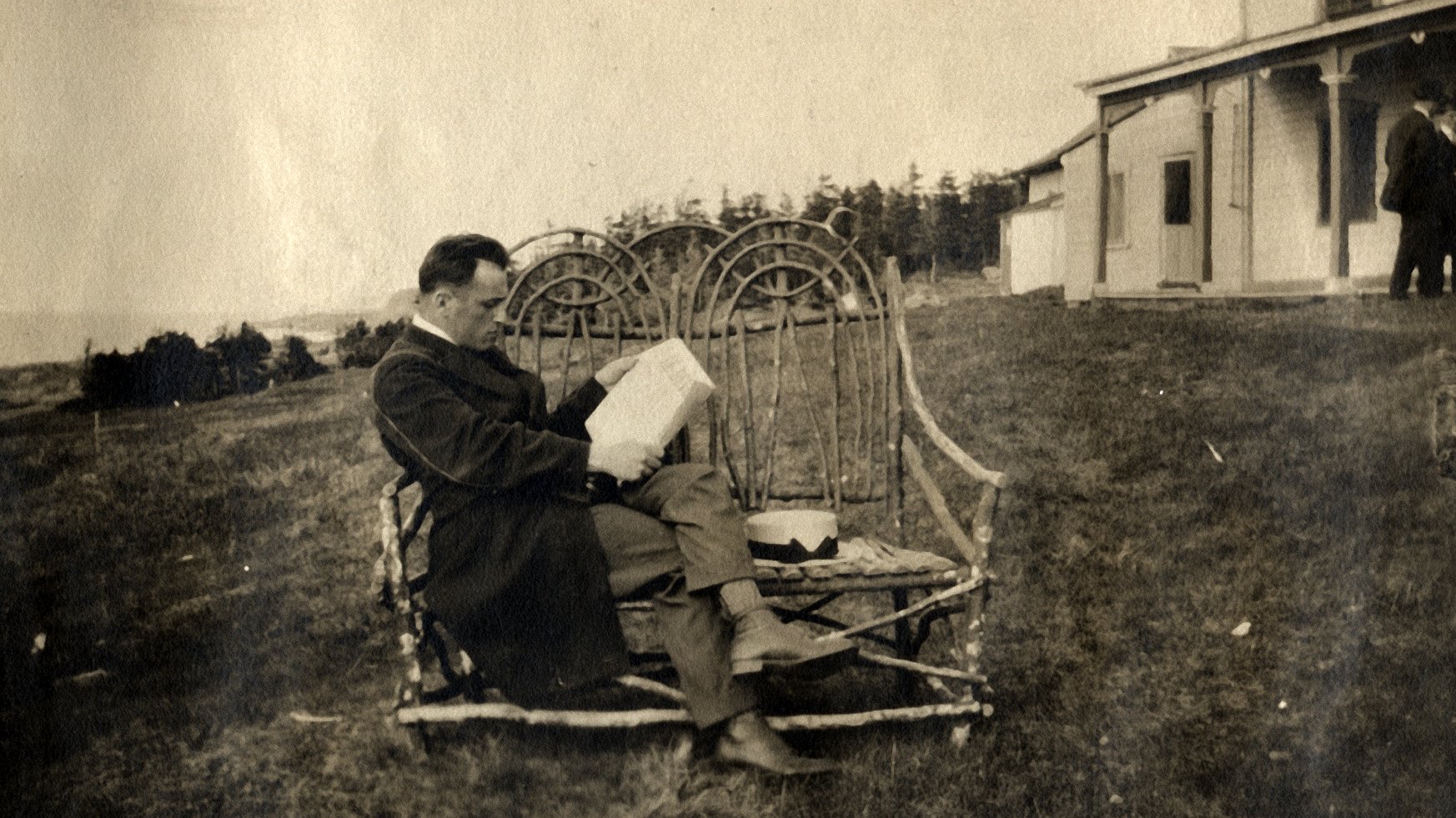 A man on vacation is reading, sitting on a wicker bench at the rear of a summer residence.