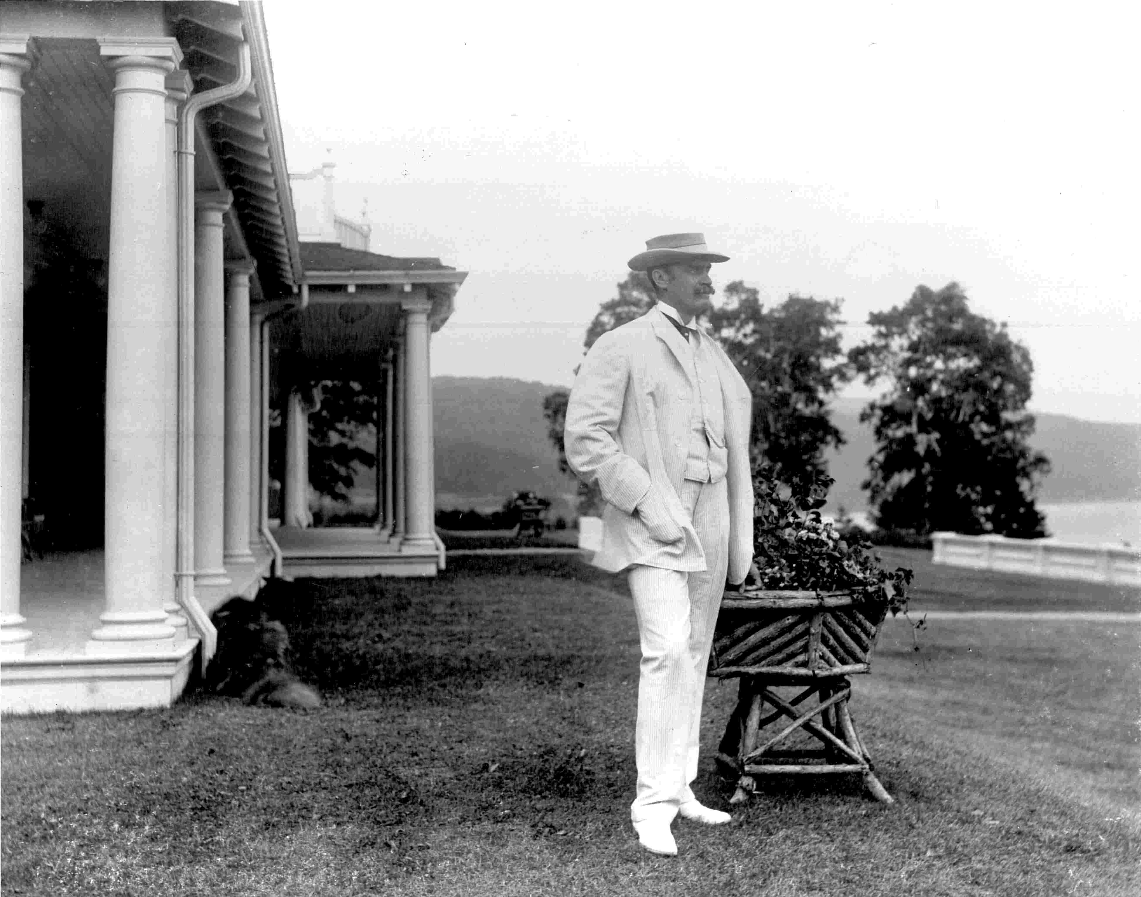 An elegant man wearing white and posing near an impressive residence, with a dog lying beside the house.