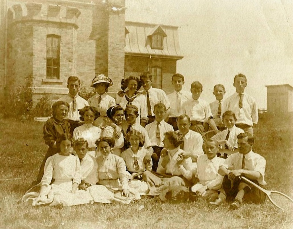 Some twenty elegantly-dressed young people pose in front of a castle-like building. A few of them hold tennis racquets.