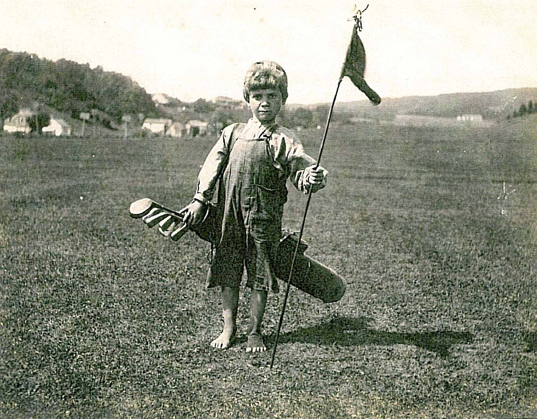 A barefoot child caddie, carrying a golf bag and holding a flag.