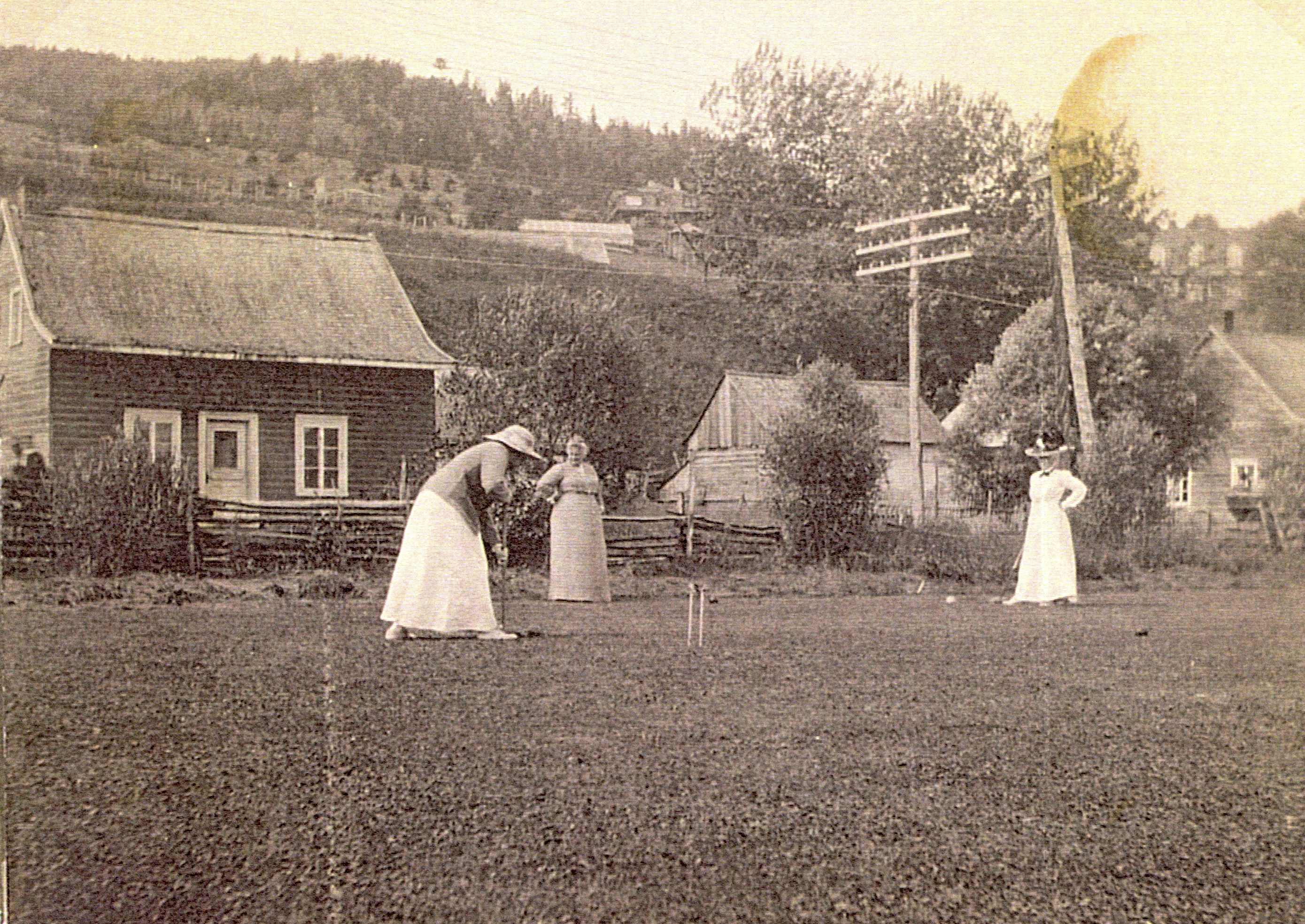 Three women play croquet near houses at the foot of a mountain.