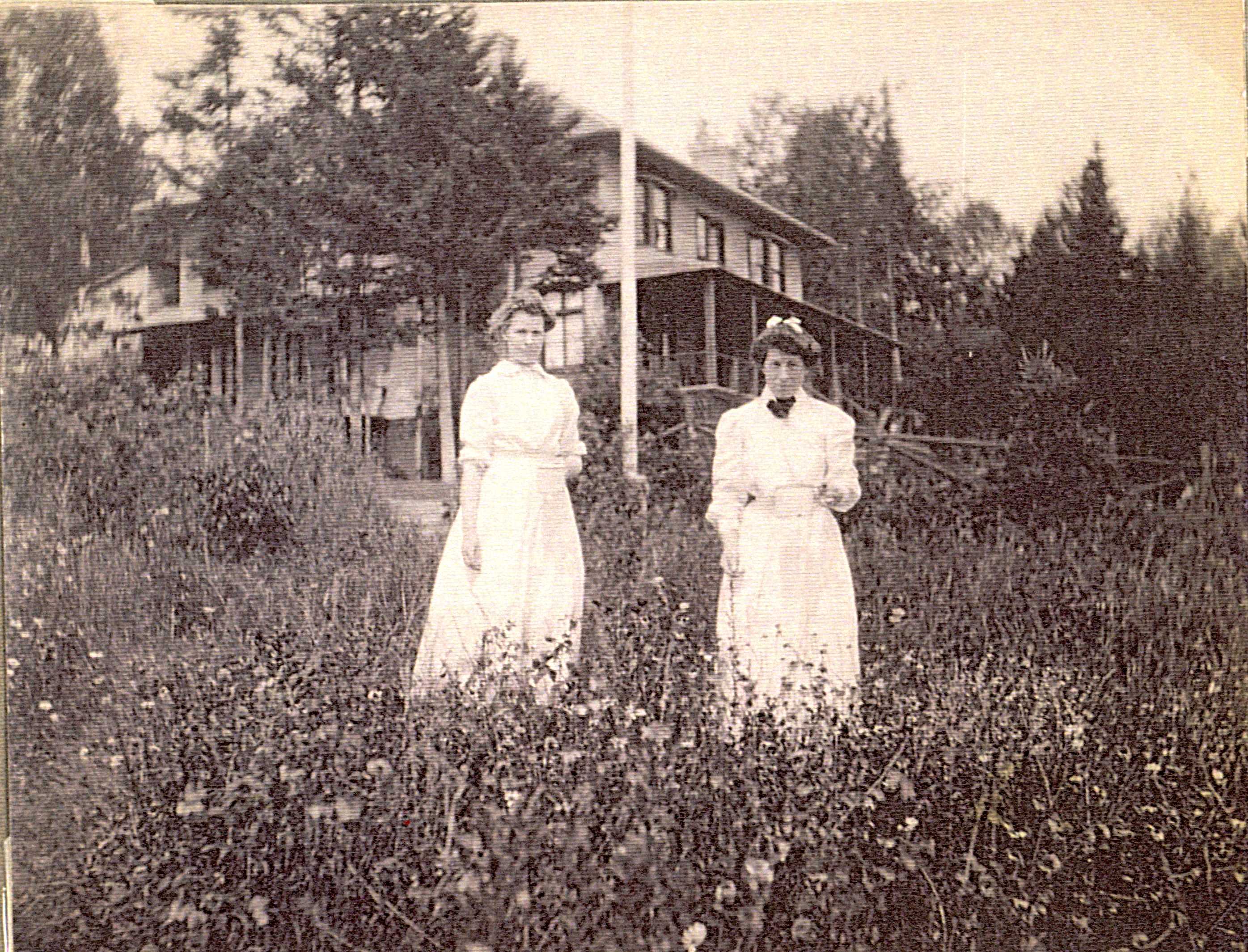 Two young women in dresses posing in a field near a house, each holding a white container.