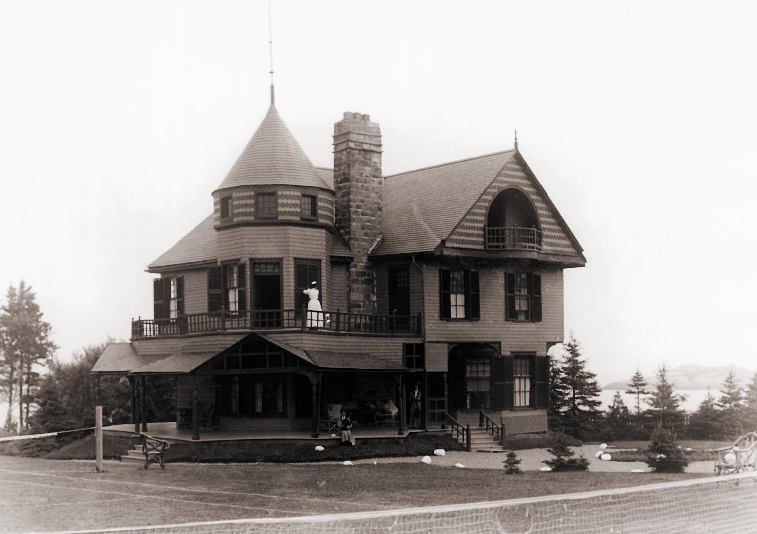 Two women are on the veranda of a luxurious house. A maid adjusts the shutters of a window on the second floor.