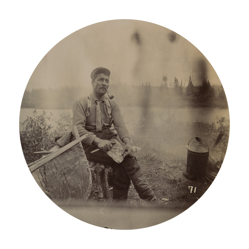 Un homme fumant la pipe est assis près d'un canot, avec un lac sauvage en arrière-plan.