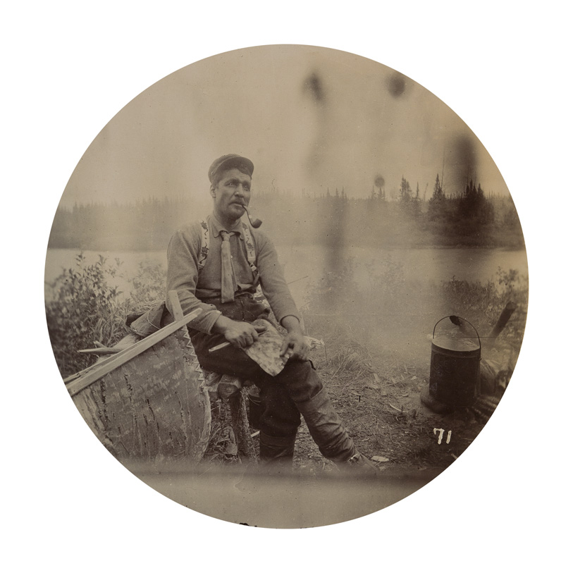 A man smoking a pipe sits by a canoe, with a lake in the background.