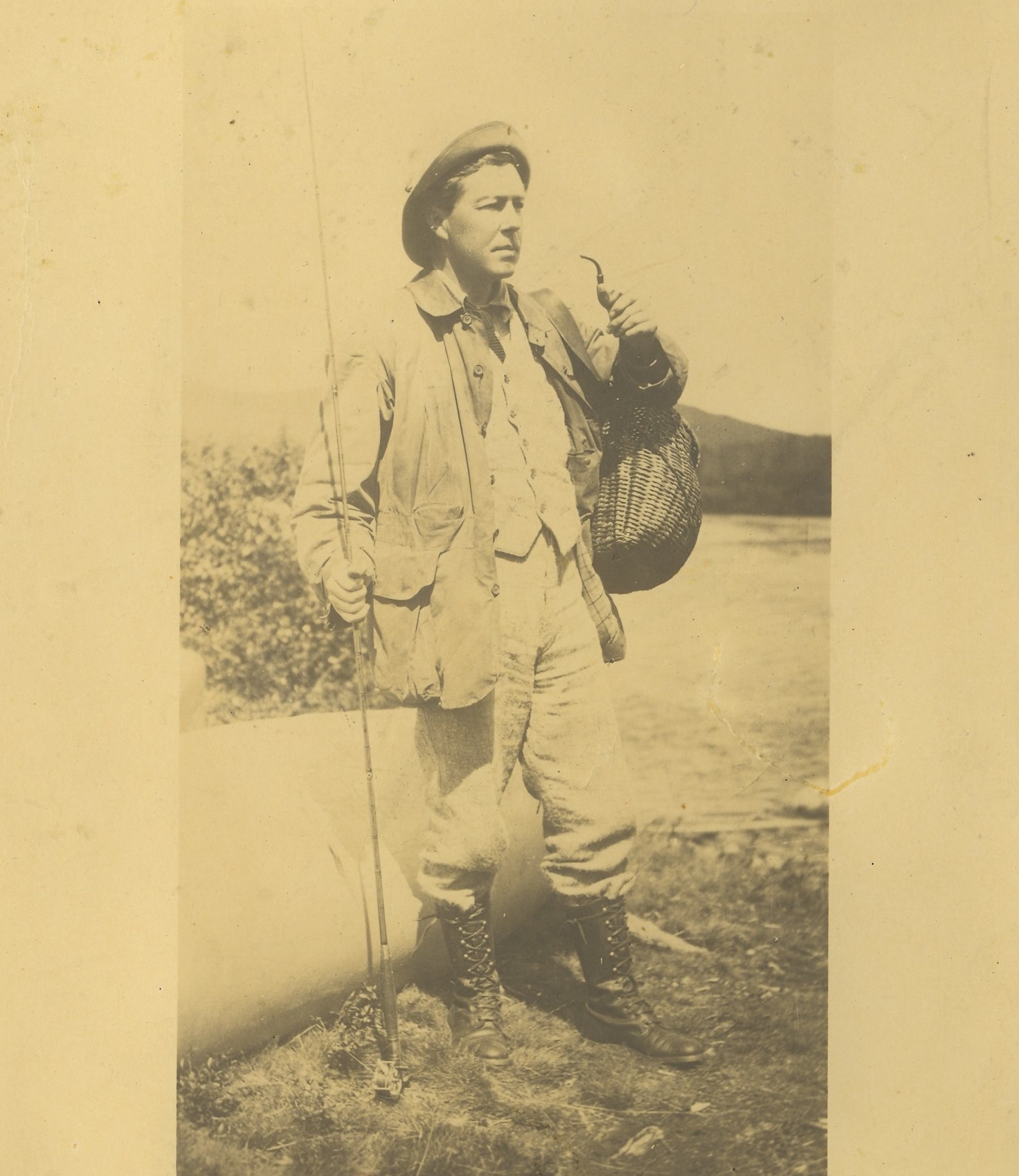 Photographie sur papier jauni d'un pêcheur et son équipement, dont une canne à pêche, un canot et panier à poissons en osier.