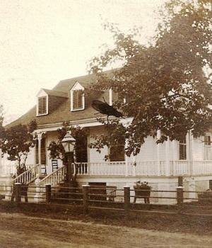 A white house with three dormer windows, located on a dirt road, is surrounded by a railed fence.