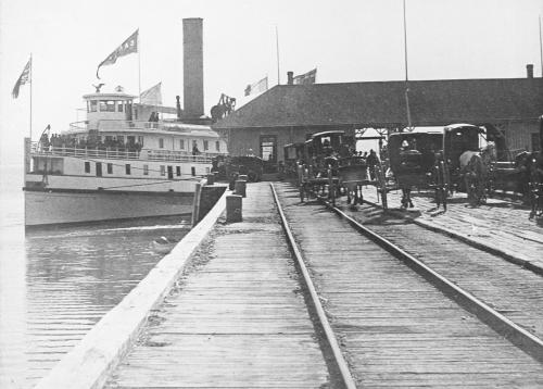 Un bateau à vapeur accosté à un quai, qui comporte une voie ferrée. Des charretiers attendent la descente des passagers.