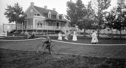 A family enjoys the summer, behind an opulent house. A child rides a bike, others play tennis or chat.