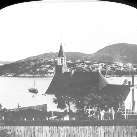 A chapel and its cemetery dominate a hill. Some houses are on the coast. A sailboat is moored.