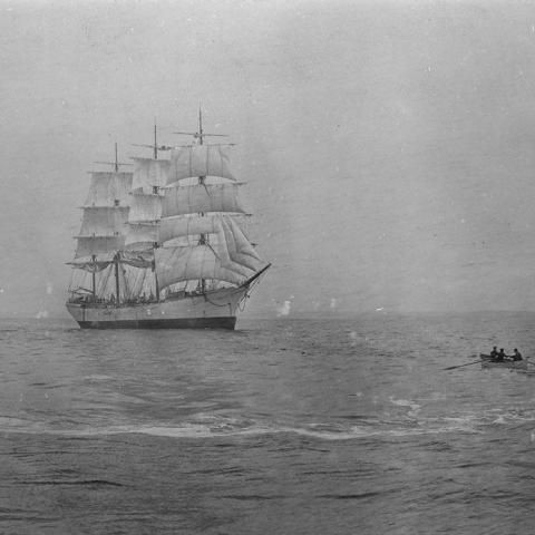 Old photography in black and white of a small boat which goes to a sailboat.