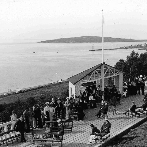 Vacationers conversing on a wood terrace built on a cliff overlooking the water.