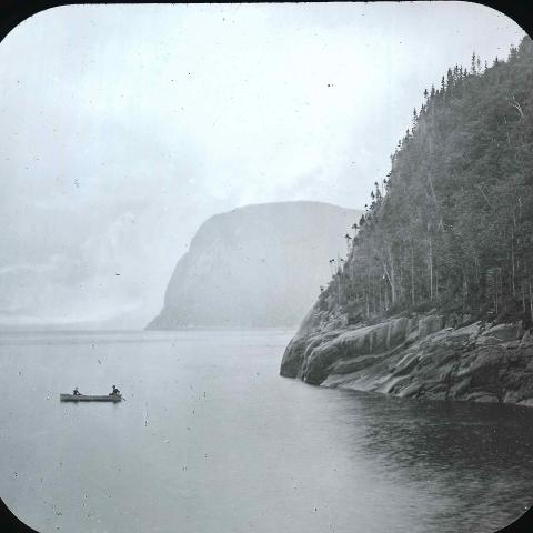 Two canoers fishingat the foot of a cliff that plunges into a very wide river.