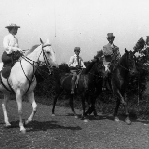 A photograph of three riders on a road: a woman mounted side-saddle, a child, and another elegant horseman.
