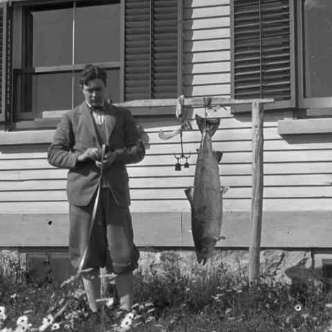 A young man rolls up a measuring tape beside an outdoor scale for weighing fish. A salmon is hanging from the scale.