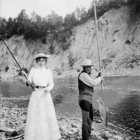 A slender woman with a fishing rod next to a guide holding a fishing net.