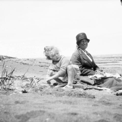 Two children play on the beach at low tide, watched over by a seated woman.