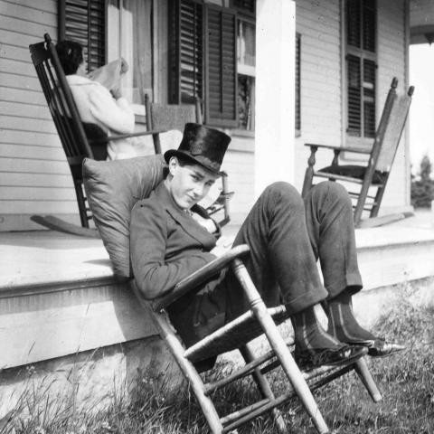 A young man wearing a top hat and sitting nonchalantly on a chair, while a woman reads in the background.