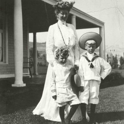 A woman in a very elegant white dress posing with her two children near a house.