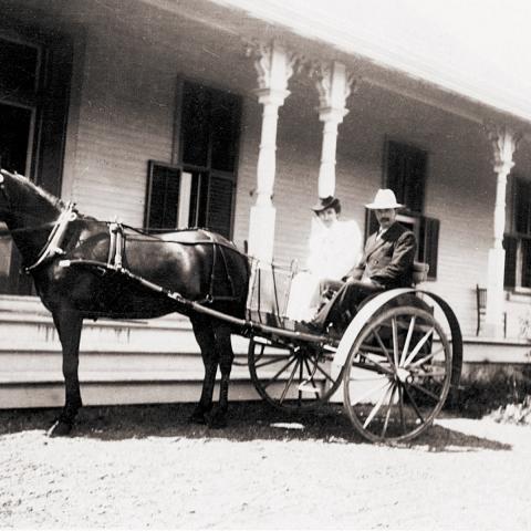 A man and a woman sitting in a calèche in front of the porch of a large building.