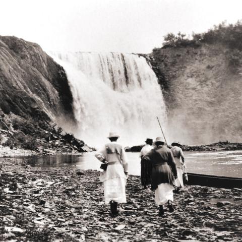 Four women walking at the foot of a waterfall, heading toward a guide standing in a canoe.