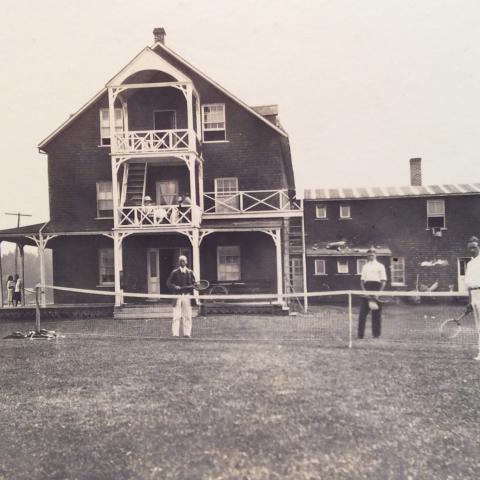 Four men play tennis on the lawn of a hotel on the shore of the St. Lawrence.