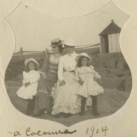 Two young women and two girls pose on rocks by the shore. A changing-cabin is in the background.