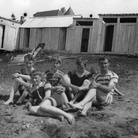 Five adolescents wearing old-fashioned bathing suits and sitting in the sand near a row of bathing cabins.