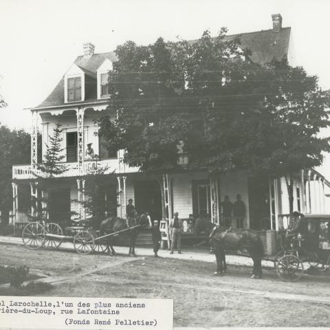 Photograph of a small hotel, with two horse-drawn carriages parked in front.