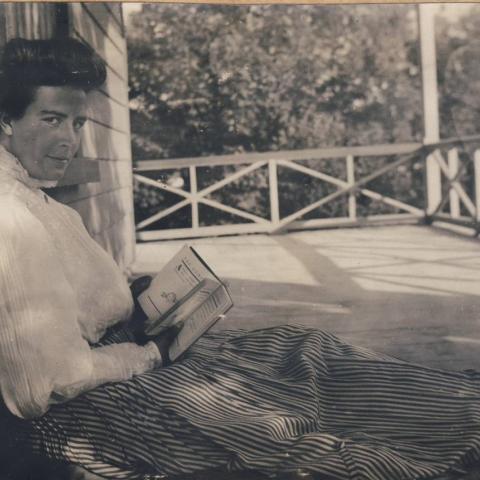 A woman reading, sitting on the floor of a broad porch, her back against the wall of the house.