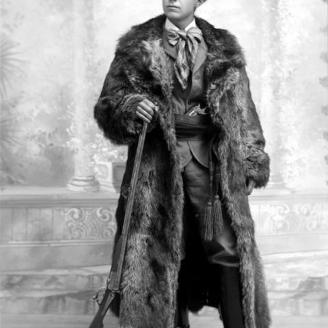 Un jeune homme vêtu d’un manteau et d’un chapeau de fourrure tient un fusil de chasse dans sa main.