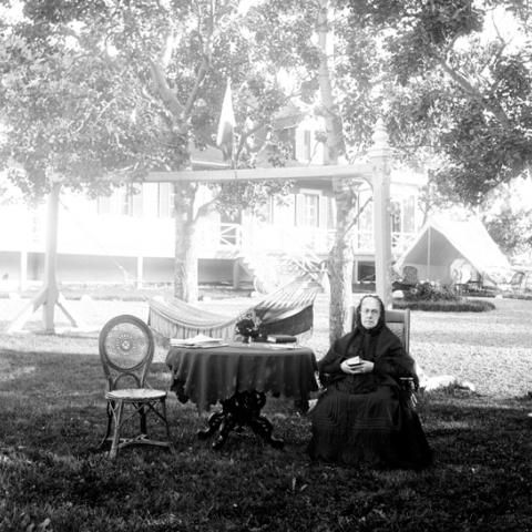 Une vieille femme lit, assise sur une chaise disposée sur le gazon.