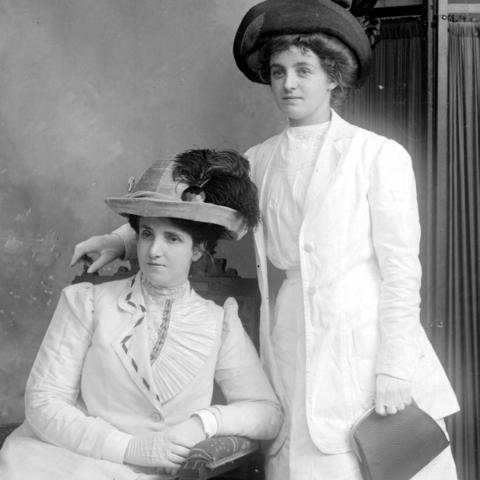 Black and white photograph of two elegant women wearing all white or very pale dresses, including gloves.