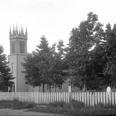 View of a small church with a neo-Gothic bell tower and connected cemetery. The lot is fenced.