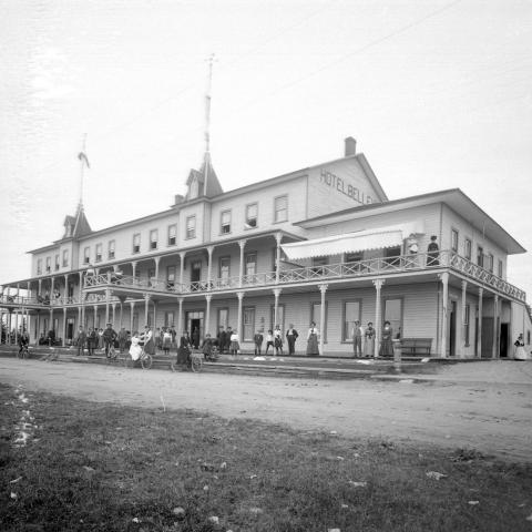 A grand wood hotel surrounded by verandas, with many tourists, including cyclists, posing on its steps.