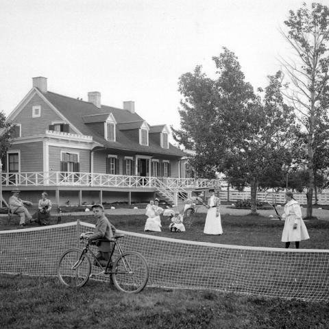 A family enjoying summer in front of a posh house and a child riding a bicycle, while others play tennis or talk.