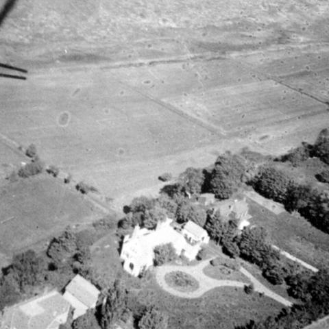 An aerial photograph in which we see a wooded estate near large fields.
