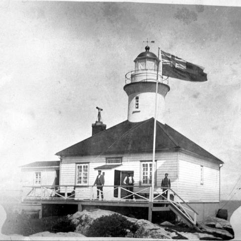 Three men pose in front of a lighthouse on the rocks. The picture is in the shape of an urn with two handles.