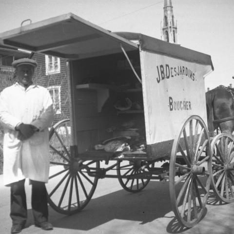 Un boucher portant un vêtement de travail blanc pose devant sa voiture de livraison à quatre roues, tirée par un cheval.