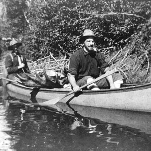 Two men in a canoe, carrying a moose.